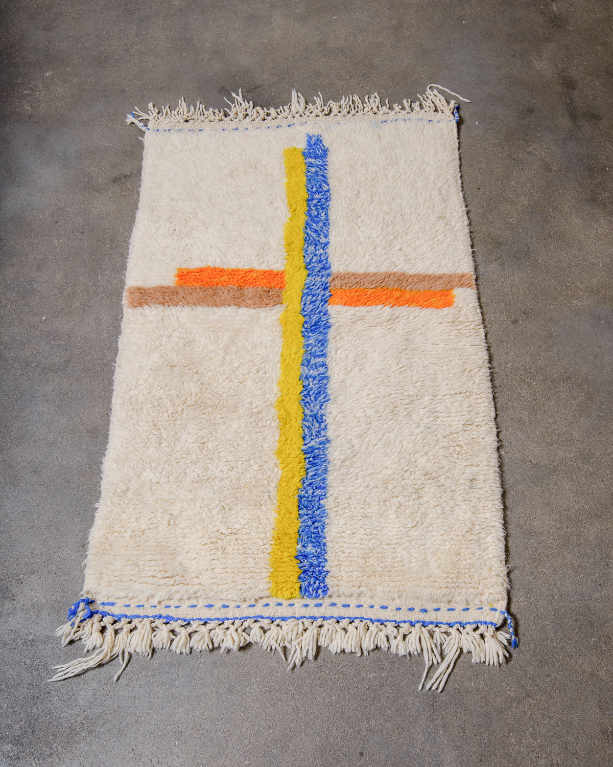 Berberlin special edition Beniourain rug made of cream wool with overlapping lines in blue, yellow, orange and brown, laying on concrete studio floor.